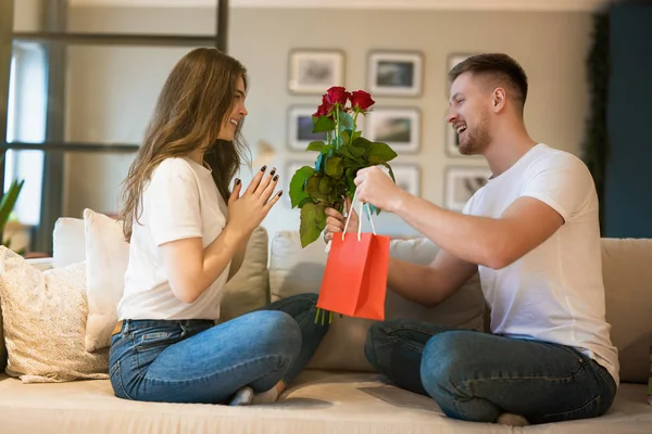 Unga vackra par sitter på soffan, man överraskande fru med blommor och gåva på helgonet Alla hjärtans dag, glad årsdag — Stockfoto