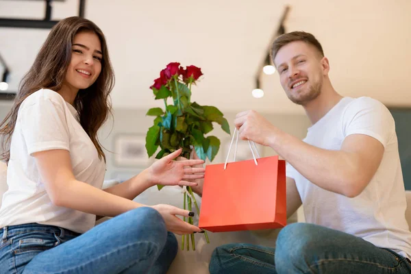 Underbart par sitter i soffan, stilig man överraskar sin vackra fru med rosor och gåva på helgonets alla hjärtans dag, glad romantisk årsdag — Stockfoto