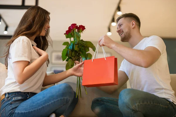 Unga vackra par sitter på soffan, man överraskande fru med rosor och gåva på helgonet Alla hjärtans dag, glad romantisk årsdag — Stockfoto