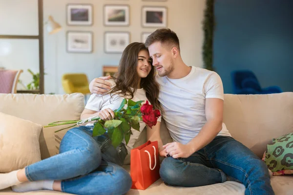 Vackra par kramas på soffan, stilig make överraska sin vackra fru med rosor och gåva på helgonet Alla hjärtans dag, glad romantisk årsdag — Stockfoto