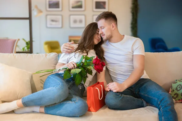 Vackra par kramas på soffan, stilig make överraska sin vackra unga fru med rosor och gåva på helgonet Alla hjärtans dag, glad romantisk årsdag — Stockfoto