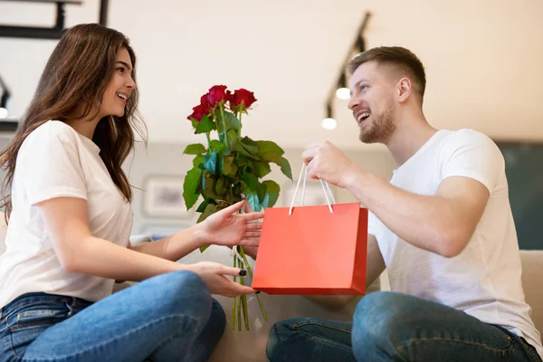 Underbart par sitter i soffan, stilig make överraskar sin vackra fru med rosor och gåva på helgonets alla hjärtans dag, glad romantisk årsdag — Stockfoto