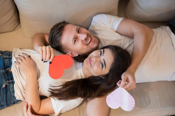 Pareja encantadora abrazándose en el sofá, marido guapo y su hermosa esposa sosteniendo corazones de papel, feliz aniversario romántico, día de San Valentín —  Fotos de Stock