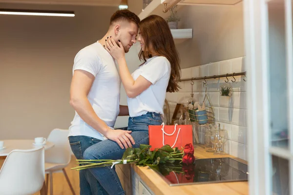 Ung stilig man överraska sin älskade fru med rosor och gåva på helgonet Alla hjärtans dag kysser henne medan hon sitter på köksbordet — Stockfoto
