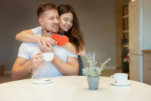 Ung vacker kvinna överraska sin man med rött papper hjärta på helgonet alla hjärtans dag under morgonen kaffe i köket, kramas, glad romantisk årsdag — Stockfoto