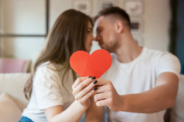 Unga par kysser på soffan, båda håller stora röda papper hörs på helgonet Alla hjärtans dag, glad årsdag — Stockfoto