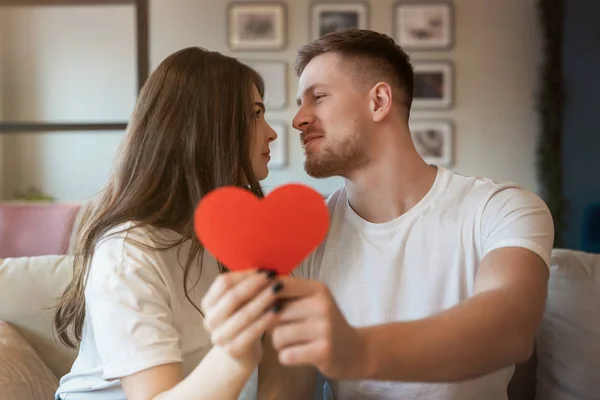 Unga romantiska par kysser på soffan, båda håller stora röda papper hörs på helgonet Alla hjärtans dag, glad årsdag — Stockfoto