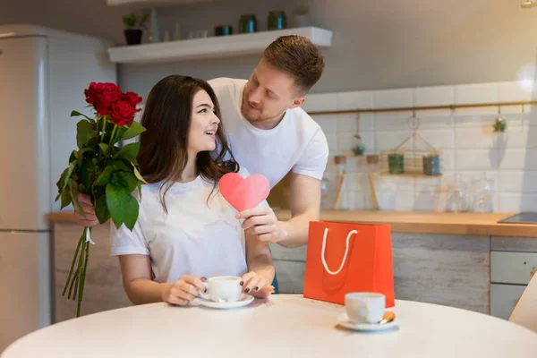 Ung stilig man överraskar sin vackra älskade fru med rosor och gåva på helgonets alla hjärtans dag på morgonen kaffe i köket, glad romantisk årsdag — Stockfoto