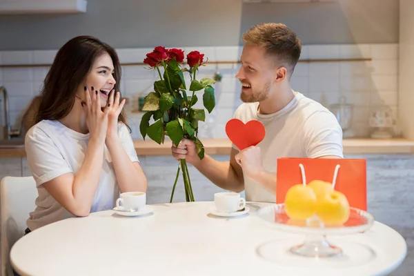 Ung stilig man överraskar sin vackra älskade fru med rosor och gåva på helgonets alla hjärtans dag under frukost, glad romantisk årsdag — Stockfoto