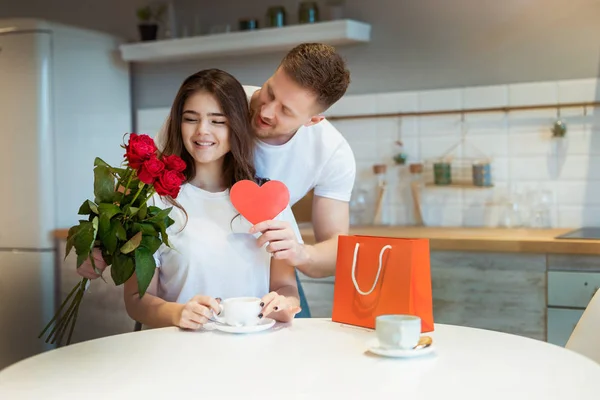 Ung vacker make överraska sin älskade fru med rosor och gåva på helgonet Alla hjärtans dag under morgonen kaffe i köket, glad romantisk årsdag — Stockfoto