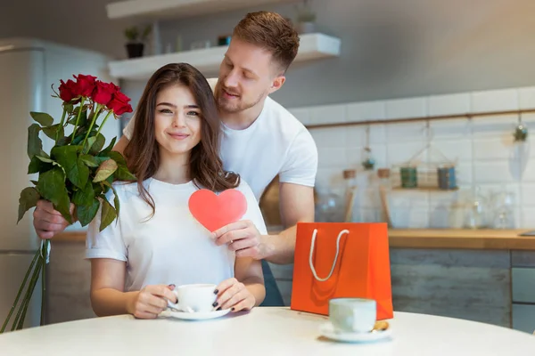 Ung stilig man överraskar sin älskade fru med rosor och gåva på helgonets alla hjärtans dag under frukost kaffe i köket, glad romantisk årsdag — Stockfoto