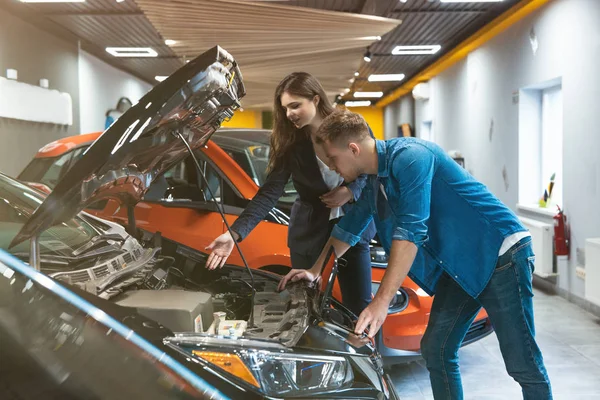 Gerente de mujer joven en traje de negocios consultando al cliente joven que muestra el motor del vehículo en el centro de concesionarios de automóviles eléctricos, cooperación profesional — Foto de Stock