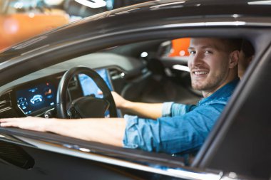 Genç yakışıklı adam yeni aracın içini kontrol ediyor. Elektrikli araba galerisinde, mutlu ve eksenli görünüyor.