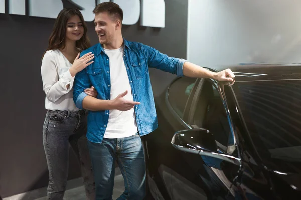 Joven feliz pareja guapo hombre y hermosa mujer comprobar nuevo interior del coche, mientras que de pie en el centro de concesionario de coches eléctricos — Foto de Stock