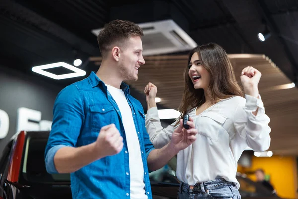 Giovane coppia di famiglia bell'uomo e bella donna cercando felice e gioioso in piedi vicino al loro veicolo nuovo di zecca nel centro di concessionaria auto elettrica, momenti indimenticabili — Foto Stock