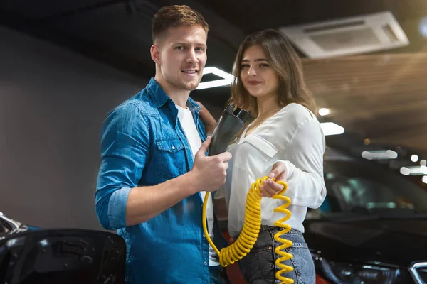 ハンサムな男と美しいブルネットの女性は電気自動車販売センターに立つ電気自動車の充電ケーブルを保持し、環境に優しい概念 — ストック写真