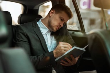 Başarılı iş adamı, ortaklarla buluşmaya giderken arabasının arka koltuğunda önemli bir telefon görüşmesi yaparken planlayıcısına notlar alır.