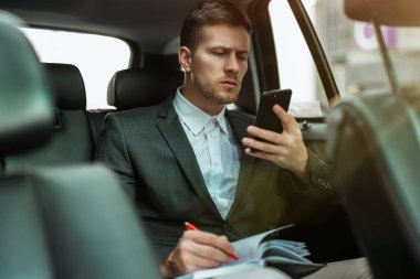 Başarılı iş adamı, ortaklarıyla buluşmaya giderken, arabasının arka koltuğunda giderken akıllı telefonundan planlayıcıya notlar alır.