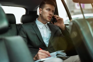 Başarılı iş adamı, ortaklarla buluşmaya giderken arabasının arka koltuğunda önemli telefon görüşmeleri yaparken planlayıcısına notlar alır.