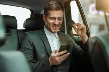 Başarılı bir işadamı, ortaklarıyla buluşmak için arabanın arka koltuğunda giderken banka kartıyla online ödeme yapıyor. Tatmin olmuş görünüyor, çoklu görev kavramı.