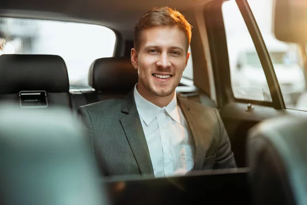 Młody szczęśliwy uśmiechnięty biznesmen pracuje w swoim laptopie siedząc w samochodzie w drodze do biura, wielozadaniowa koncepcja — Zdjęcie stockowe