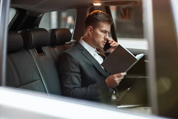 Молодой бизнесмен, беседующий по телефону, держит своего планировщика, сидя в машине на пути к встрече с партнерами, многозадачная концепция — стоковое фото
