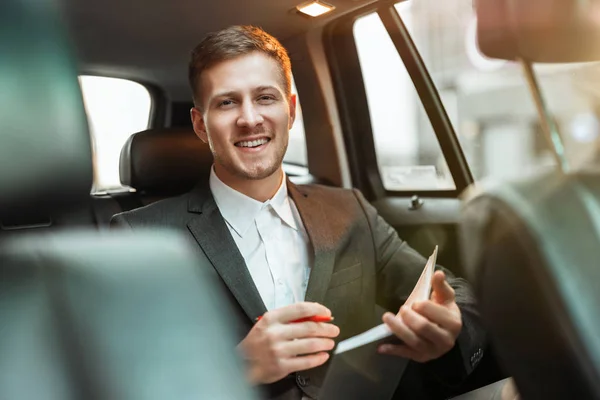 Młody odnoszący sukcesy biznesmen robi notatki swojemu planiście siedząc w samochodzie w drodze na spotkanie z partnerami, wygląda na szczęśliwego, wielozadaniową koncepcję — Zdjęcie stockowe
