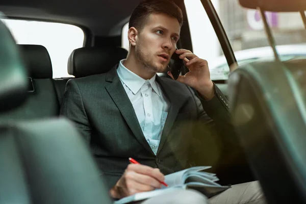 Успешный бизнесмен делает заметки к планировщику, имея важные телефонные переговоры езда на заднем сиденье автомобиля на пути к встрече с партнерами, многозадачность концепции — стоковое фото