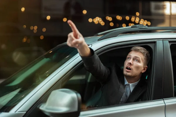 Młody biznesmen wskazując palcem z otwartego okna samochodu, wygląda zdumiewająco, duże życie miasta — Zdjęcie stockowe