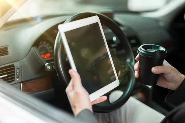 Uomo d'affari tiene il caffè in una tazza di papper e legge notizie dal suo tablet mentre seduto in macchina con finestra aperta, multitasking, grande vita di città — Foto Stock