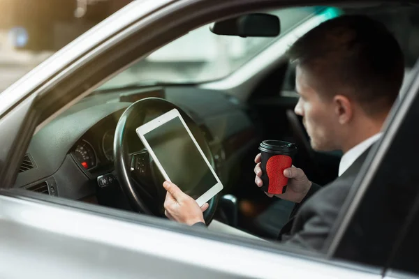 Giovane uomo d'affari beve caffè e legge notizie dal suo tablet mentre è seduto in macchina con finestra aperta, multitasking, grande vita di città — Foto Stock