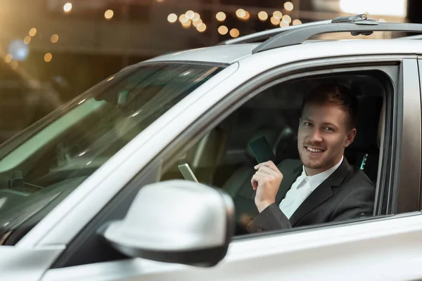 Młody uśmiechnięty biznesmen wygląda szczęśliwy podczas jazdy samochodem z otwartym oknem, wielozadaniowość, big city life — Zdjęcie stockowe
