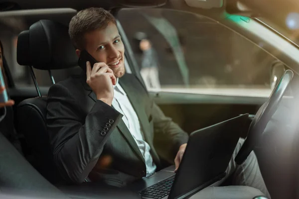 Młody uśmiechnięty biznesmen pracuje w swoim samochodzie, korzystając z laptopa, rozmawiając na smartfonie przygotowując się do ważnego spotkania z partnerami, wielozadaniowa koncepcja — Zdjęcie stockowe