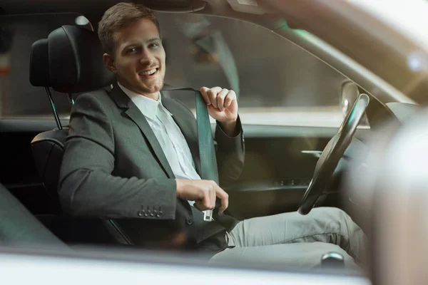Молодой улыбающийся бизнесмен сидит в своей машине выглядит счастливым и удовлетворенным, регулируя свой ремень безопасности перед началом поездки, концепция безопасности вождения — стоковое фото