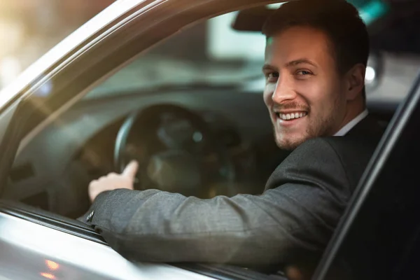 Молодой улыбчивый успешный бизнесмен выглядит счастливым за рулем своей машины с открытым окном, многозадачной, большой городской жизнью — стоковое фото