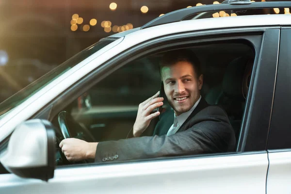 Молодой улыбающийся успешный бизнесмен с приятным телефонным разговором за рулем своей машины с открытым окном, многозадачность, большая городская жизнь, безопасное вождение — стоковое фото
