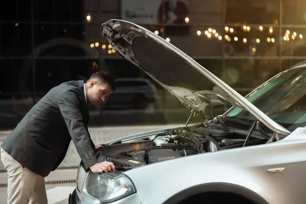 Joven hombre de negocios guapo de pie cerca de parachoques de coche delantero abierto, comprobar el motor, se ve molesto, problemas inesperados del vehículo — Foto de Stock