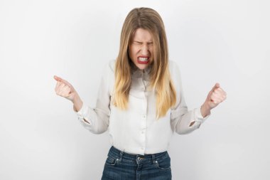 Genç, sarışın, kızgın kadın öfkeli görünüyor. Yumruklarını sıkmış, izole edilmiş beyaz arka planda duruyor. Duygularını belli ediyor.