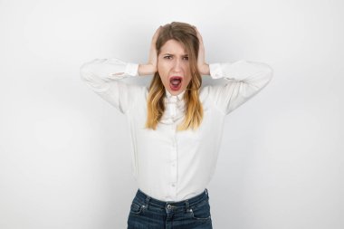 Genç ve güzel sarışın bir kadın, gözleri tamamen açık, izole beyaz arka planda duruyor, iki eliyle kulaklarını kapatıyor, yüksek sesle bağırıyor, korkmuş görünüyor.
