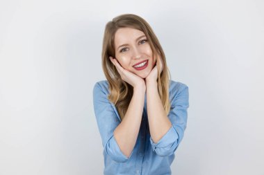 Genç, güzel, sarışın, gülümseyen bir kadın iki eliyle yüzüne dokunarak tatmin olmuş görünüyor. İzole edilmiş beyaz arka plan, vücut dili.