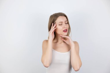 Genç ve güzel sarışın kadın, teninden memnun görünüyor. Yüzüne dokunuyor. İzole edilmiş beyaz arka plan, sağlık sigortası ve güzellik kavramına kapalı gözlerle bakıyor.