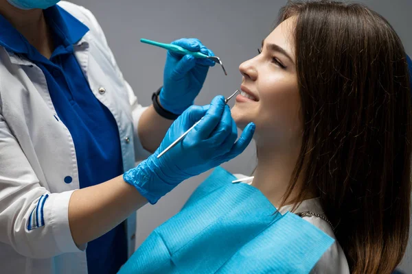 Dentista con guantes azules examina sonriente paciente usando escamadora dental mientras ella está sentada medio turno en la silla dental en la clínica, concepto de salud — Foto de Stock