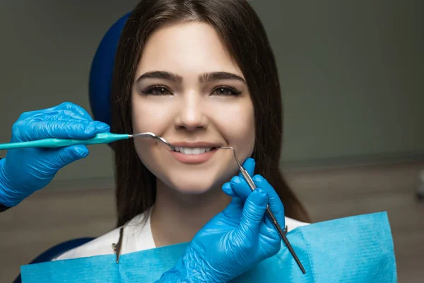 Uśmiechnięta piękna szczęśliwa brunetka pacjentka przebadana przez dentystę w niebieskich rękawiczkach za pomocą lusterka dentystycznego i skalera podczas wizyty w klinice stomatologicznej, koncepcja opieki zdrowotnej — Zdjęcie stockowe