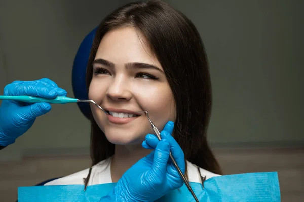 Patiente brune examinée par un dentiste en gants bleus à l'aide d'un miroir dentaire et d'un détartreur assis une demi-tour dans une chaise dentaire pendant le rendez-vous, concept de soins de santé — Photo