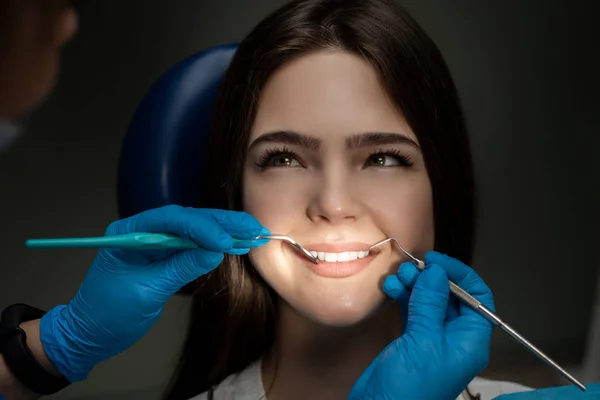 Szczęśliwy brunetka pacjent badane przez dentysta w niebieskich rękawiczkach za pomocą przyrządów dentystycznych siedzi w fotelu stomatologicznym pod lampą w klinice, zbliżenie — Zdjęcie stockowe