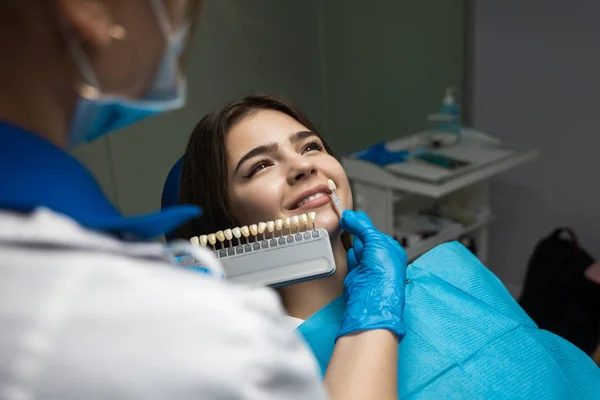 Stomatolog w niebieskich rękawiczkach medycznych i masce nakładając próbkę ze skali emalii zęba na zęby pacjentki, aby podnieść odpowiedni odcień do zabiegu wybielania — Zdjęcie stockowe