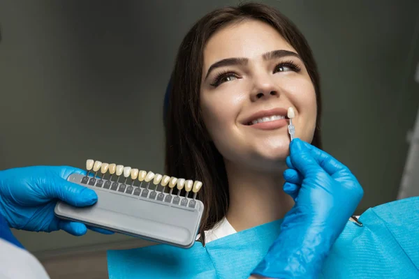 Dentysta w niebieskich rękawicach medycznych stosujących próbkę z emaliowanej skali zębów do szczęśliwej kobiety zębów pacjenta, aby odebrać prawy odcień dla procedury wybielania zębów — Zdjęcie stockowe