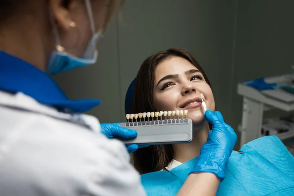 Dentista en guantes médicos azules y máscara que aplica la muestra de la escala de esmalte dental a los dientes del paciente de la mujer para elegir la sombra correcta para el procedimiento de blanqueo — Foto de Stock