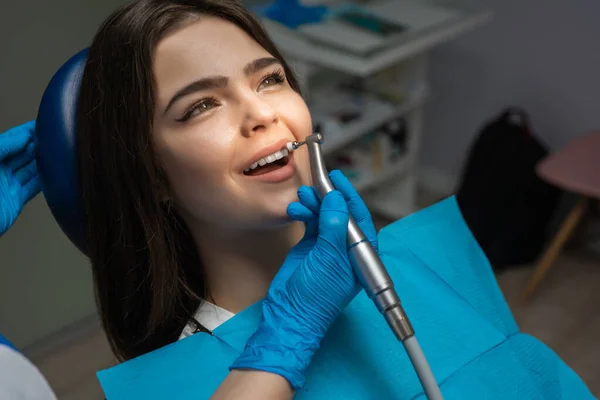 Brunetka žena pacient vyšetřen zubař v modrých rukavicích pomocí zubní zrcadlo a škálovač sedí v zubním křesle během vytvrzování zubů na klinice, helthcare koncept — Stock fotografie