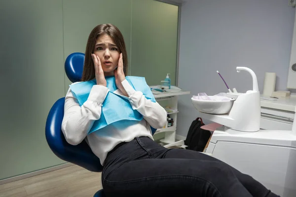 Belle jeune femme patiente effrayée assise dans une chaise dentaire pendant l'examen au cabinet dentaire toucher désespérément son visage semble effrayé, concept de soins de santé — Photo